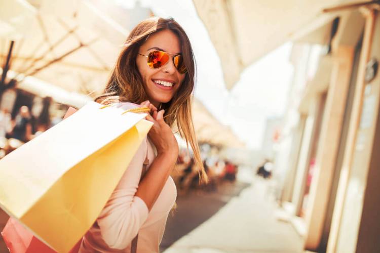 Woman in aviator sunglasses looking over her shoulder holding a yellow gift bag in a sunlit mall