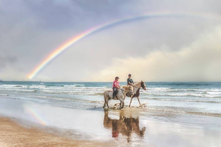 Two people riding horses on the beach with a rainbow in the background over the ocean